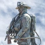 Bronze statue of a firefighter in uniform holding an axe, with hoses and equipment on his back, set against a cloudy sky background.