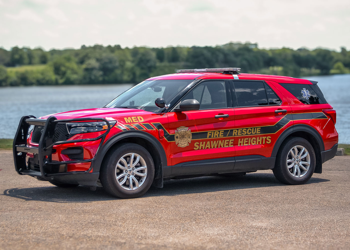 A bright red Shawnee Heights Fire/Rescue SUV is parked on pavement near a body of water, with trees and greenery visible in the background. The vehicle has emergency markings and a push bumper.