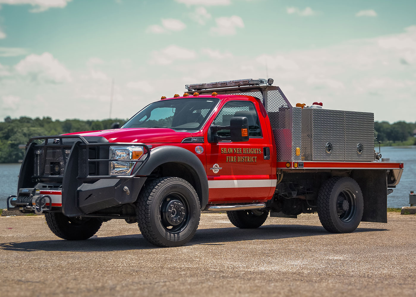 A red Shawnee Heights Fire District truck is parked outdoors on pavement near a body of water, with trees and a partly cloudy sky in the background.