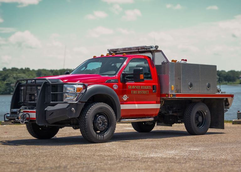 A red Shawnee Heights Fire District truck with a black front bumper and metal side compartments is parked on a paved surface near water and trees under a partly cloudy sky.