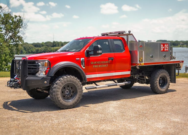 A bright red fire department rescue truck labeled "Shawnee Heights Fire District B-25" is parked outdoors on gravel, with trees and a lake in the background under a clear sky.