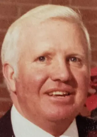 An older man with short white hair is smiling. He is wearing a white shirt and dark jacket. The background features a brick wall.