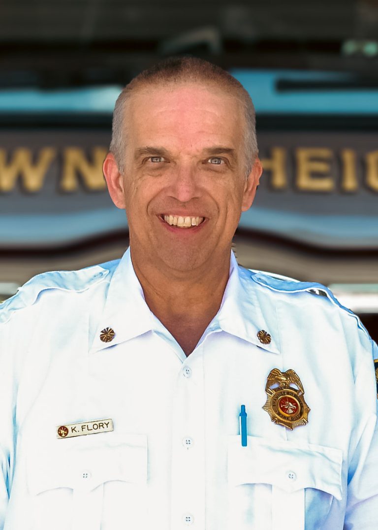 A smiling man in a white fire department uniform with badges and a name tag stands in front of a fire truck.