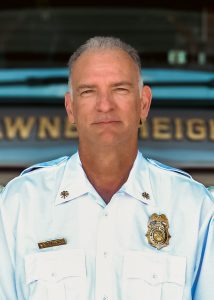 A middle-aged man in a light blue uniform shirt stands in front of a vehicle with gold lettering. He has short gray hair and wears a badge and nametag on his shirt, suggesting he is a fire or police chief.