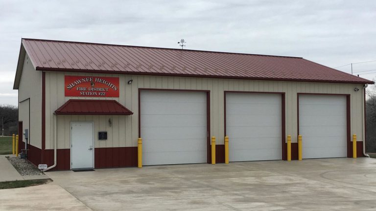 A beige and red metal building with a red roof and four large garage doors, labeled "Shawnee Heights Fire District Station #22" above the entrance, sits on a paved lot under a cloudy sky.