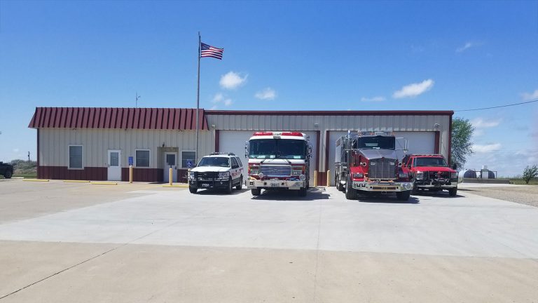 A small fire station with a U.S. flag flying in front, featuring two fire trucks and two emergency vehicles parked outside under a clear blue sky.