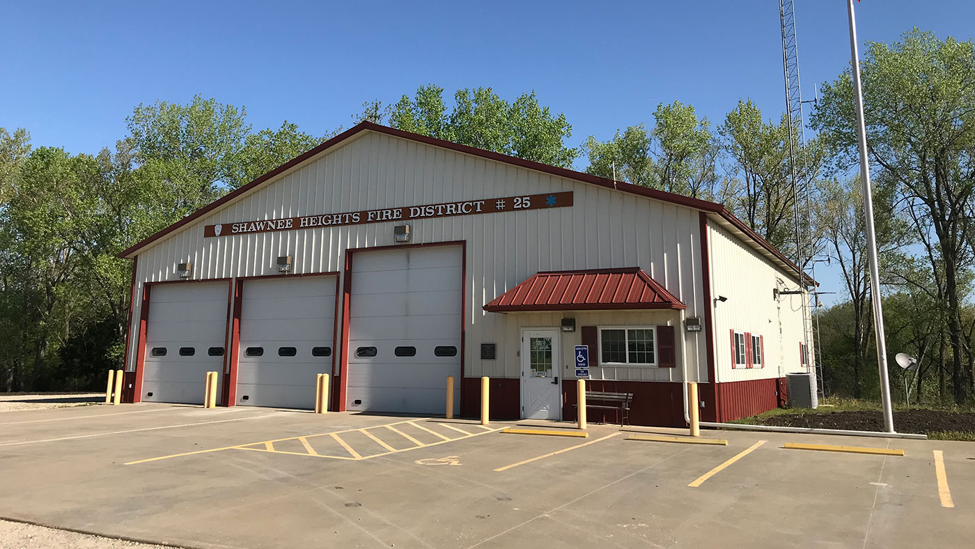 A beige and red fire station building with three large garage doors, labeled “Shawnee Heights Fire District #25.” There is a small entrance with a ramp and accessible parking in front, surrounded by trees.
