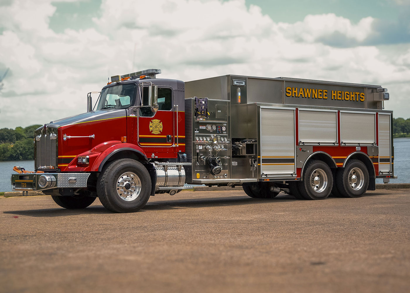A red and silver fire truck with "SHAWNEE HEIGHTS" written on the side is parked on a paved surface near a body of water, with trees and a cloudy sky in the background.