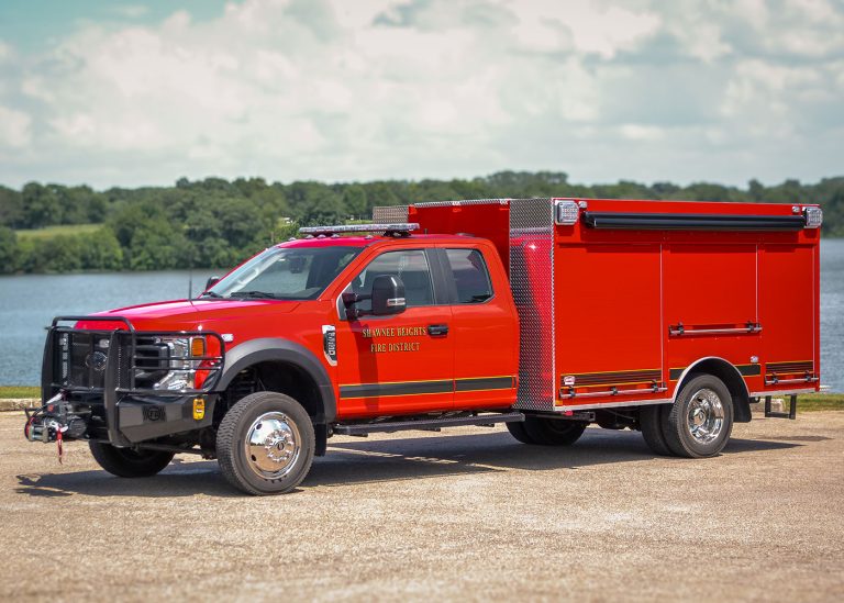 A bright red fire rescue truck is parked on a gravel road beside a lake, with trees and greenery in the background under a partly cloudy sky.