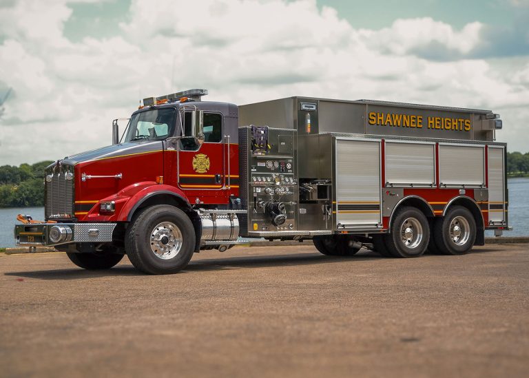 A large red and silver Shawnee Heights fire truck with chrome accents is parked on a paved surface near water, under a partly cloudy sky.