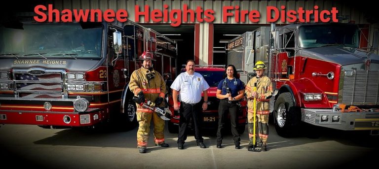 Four firefighters in uniform stand in front of two fire trucks and a fire SUV, with "Shawnee Heights Fire District" written in large red letters above them.