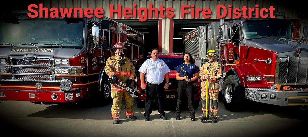 Four firefighters in uniform stand in front of two fire trucks and a fire SUV, with "Shawnee Heights Fire District" written in large red letters above them.