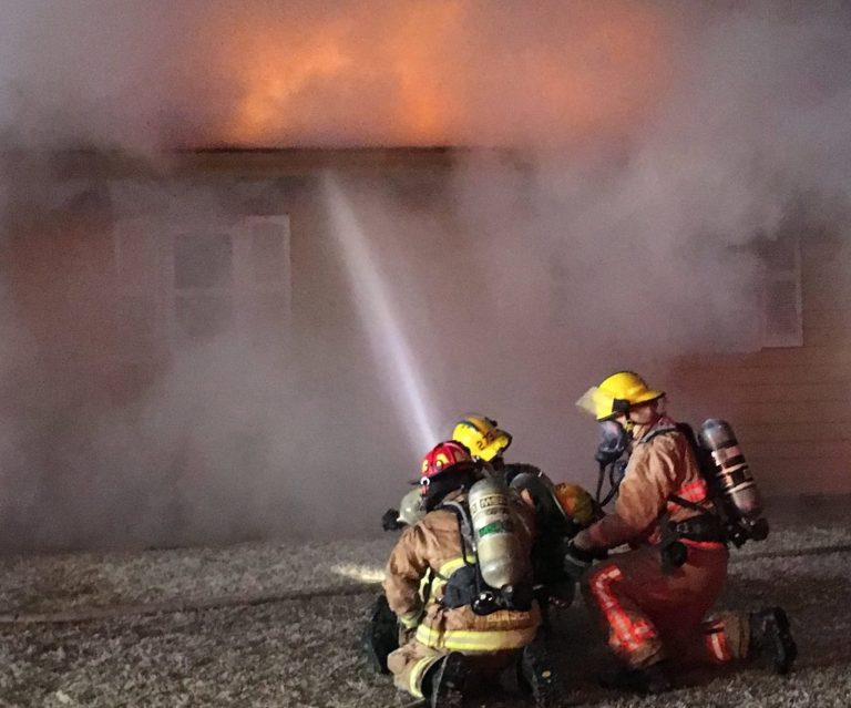 Three firefighters in full gear kneel on the ground, spraying water at a house engulfed in thick smoke and orange flames coming from the roof.