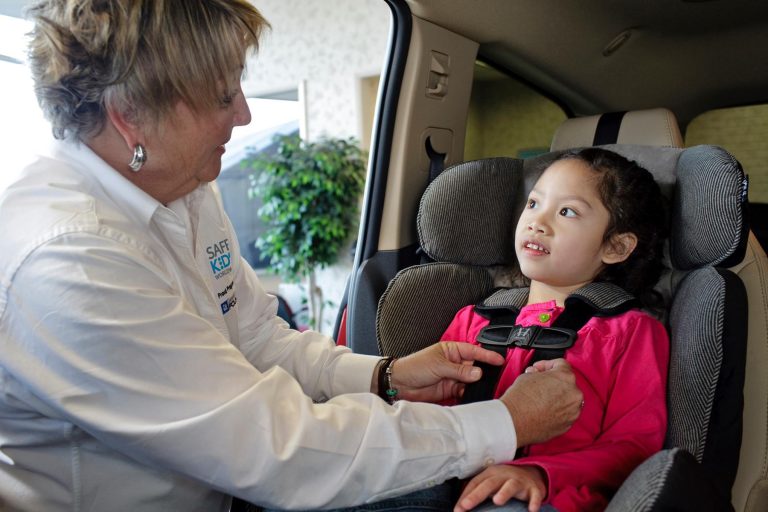 An adult helps a young girl secure her seatbelt in a car seat inside a vehicle. The woman adjusts the straps while the child, wearing a pink jacket, looks up and smiles.