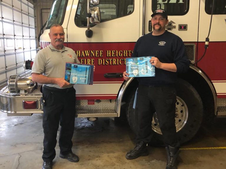 Two firefighters stand in front of a fire truck labeled “Shawnee Heights Fire District,” each holding certificates and smiling inside a fire station garage.