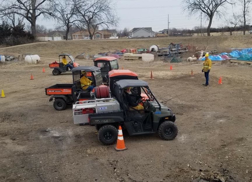 Three utility vehicles with firefighting equipment and workers in yellow jackets are parked on a dirt lot, surrounded by traffic cones. One person stands nearby, and the background has various supplies and bare trees.