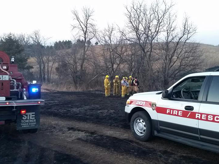 Several firefighters in yellow gear stand near burned grass and leafless trees, while a fire rescue SUV and emergency vehicle with flashing lights are parked nearby on a rural road.