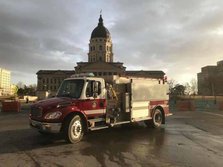 A red and white fire truck is parked in front of a large, domed government building under a cloudy sky, with sunlight breaking through.