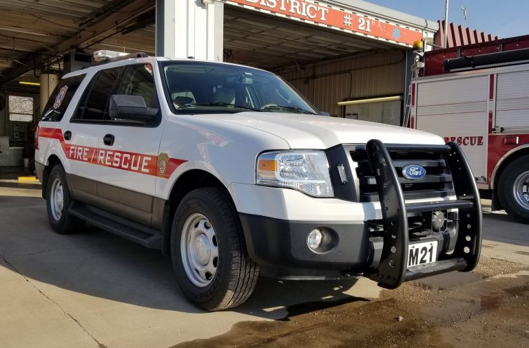 A white Ford SUV marked "FIRE/RESCUE" is parked outside a fire station, equipped with emergency lights and a push bumper. Part of a fire truck is visible in the background.