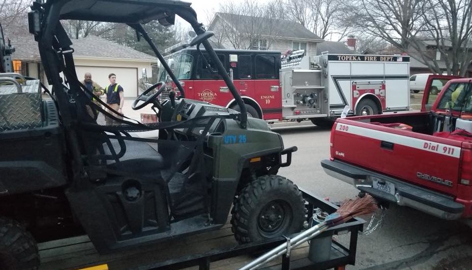 A utility vehicle and a red pickup truck with a broken rear window are parked on a residential street. A fire truck and several firefighters are in the background near houses.