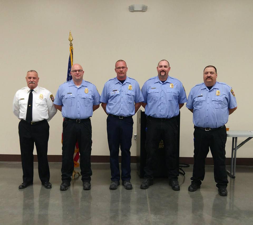 Five firefighters stand indoors in a row, four in light blue uniforms and one in a white shirt and black tie, with an American flag behind them and a table on the right.