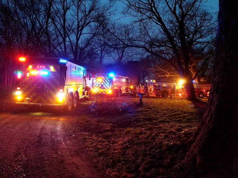 Several fire trucks with flashing lights are parked along a wooded dirt road at dusk. Firefighters are gathered near the vehicles under bare trees, with illuminated emergency lights reflecting off the scene.