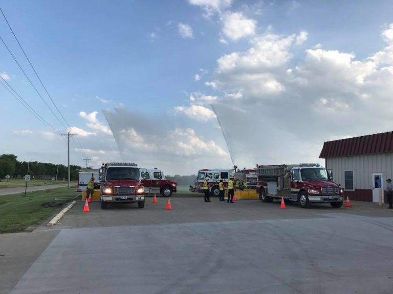 Several fire trucks are parked outside a building with firefighters standing nearby. Water streams arc into the sky, and orange cones are set up on the pavement. The sky is partly cloudy.