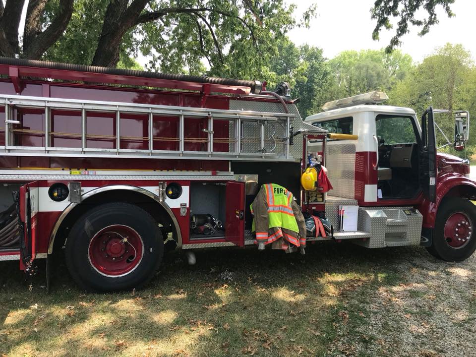 A red fire truck is parked on grass under trees. Firefighting gear, including a helmet and a reflective jacket labeled "FIRE," hangs on the side next to an open cab door. Sunlight filters through the leaves above.