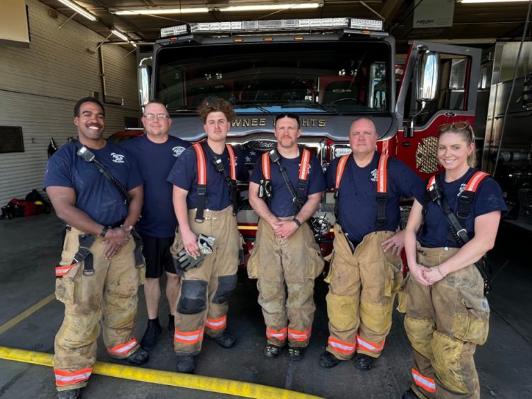 Six firefighters in uniform and turnout gear stand smiling in front of a large fire truck inside a fire station. The fire truck reads “SHAWNEE HEIGHTS” across the front.