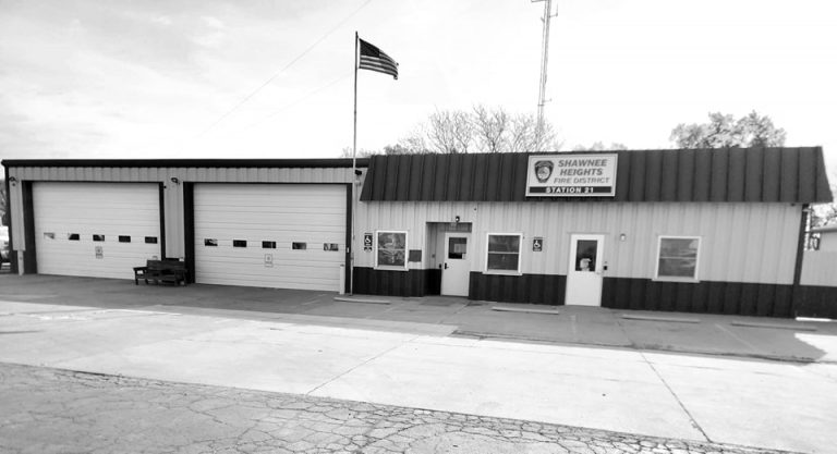 A black and white image of a fire station building with two large garage doors, an American flag on a pole, and a sign that reads "Shawnee Heights Fire District Station 21." The area is paved with concrete.