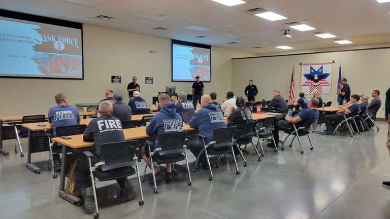 A group of firefighters in uniform sit at tables in a classroom, facing presenters and a screen displaying "TASK FORCE 0." An American flag with a firefighter emblem hangs on the wall.
