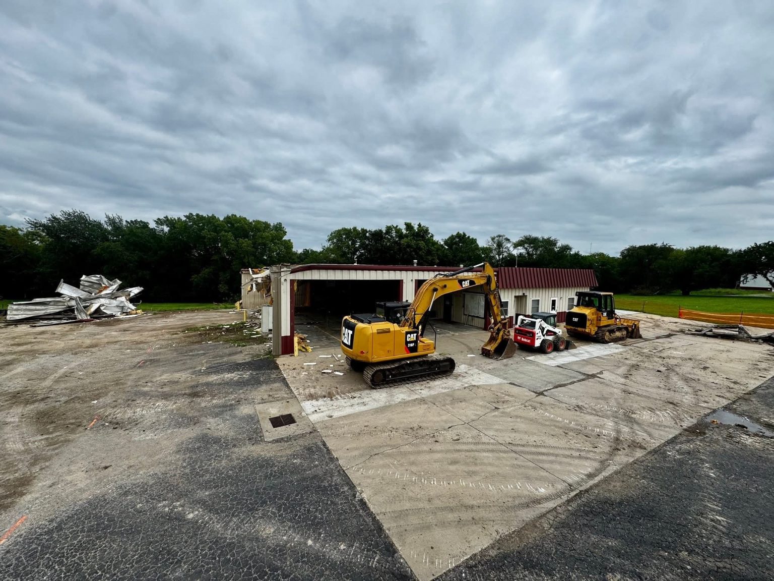 A yellow excavator and a bulldozer are parked in front of a partially demolished building, with debris piled to the left and cloudy skies overhead. Trees and grassy areas surround the construction site.