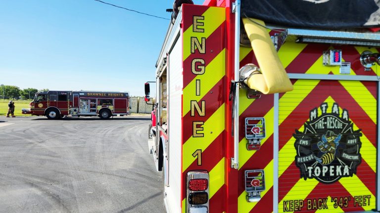 Two fire trucks are parked on a paved road. The nearest one is marked "Engine 1" with bright red and yellow stripes and "Topeka Fire-Rescue" on the back. Another fire truck is in the distance on the left.