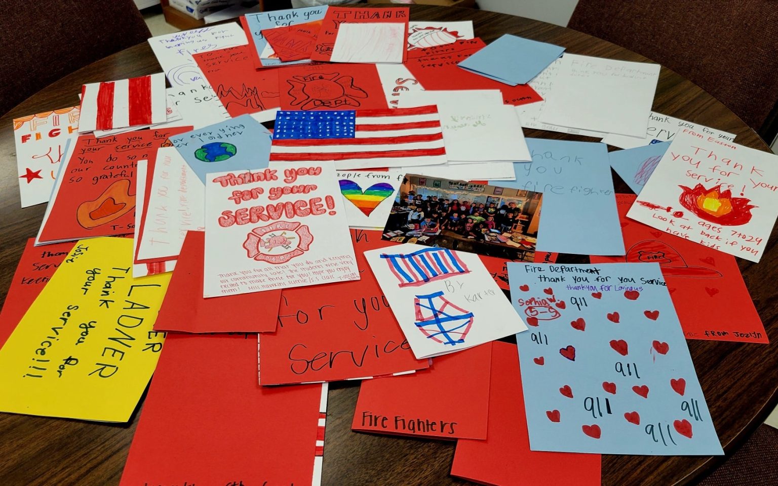 A table covered with colorful thank-you cards and drawings made by children, many featuring fire trucks, American flags, hearts, and messages expressing gratitude for firefighters' service.