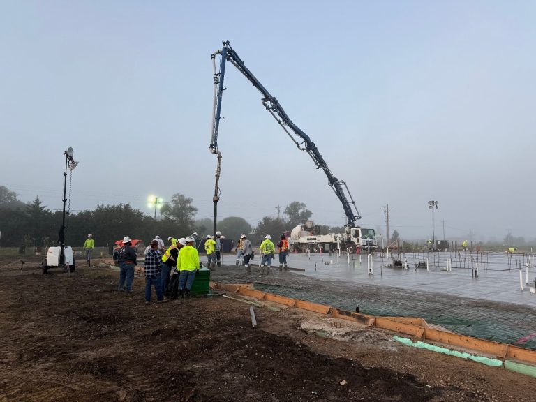 Construction workers in safety gear gather at a building site while a large concrete pump truck pours concrete onto the foundation. The scene is misty, with construction materials and equipment visible around the site.