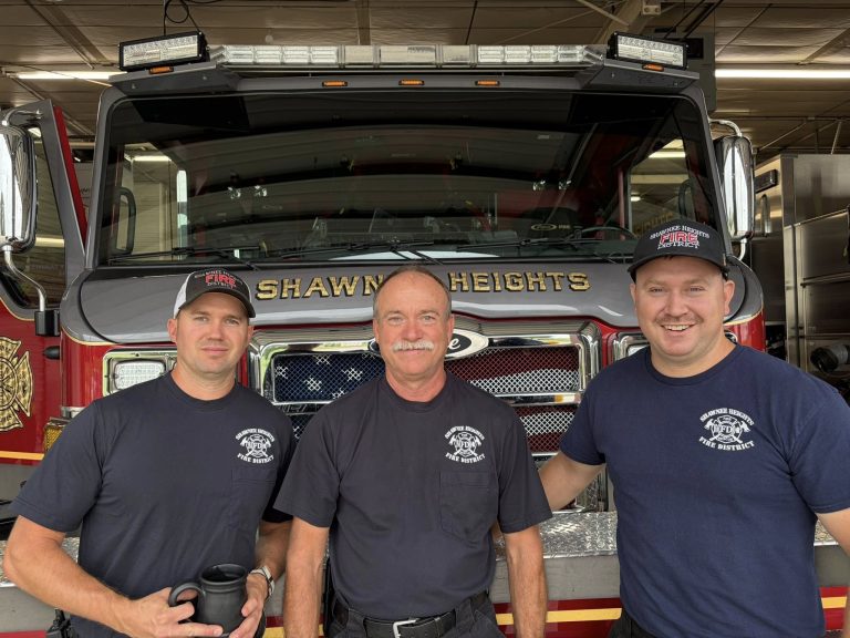 Three firefighters in navy shirts and hats stand smiling in front of a red fire truck labeled "Shawnee Heights," inside a fire station.