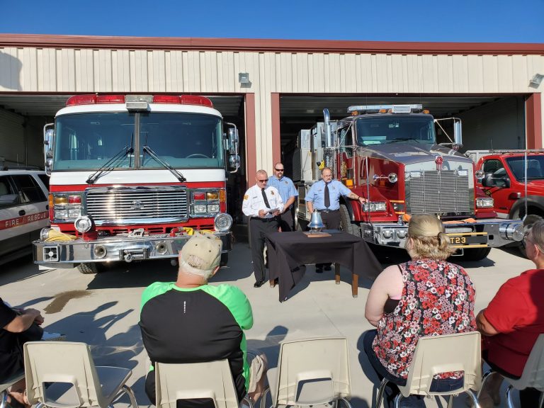 A group of people sit facing two fire trucks parked in front of a fire station. Three uniformed firefighters stand at a table with a bell and papers, appearing to hold a ceremony or presentation.