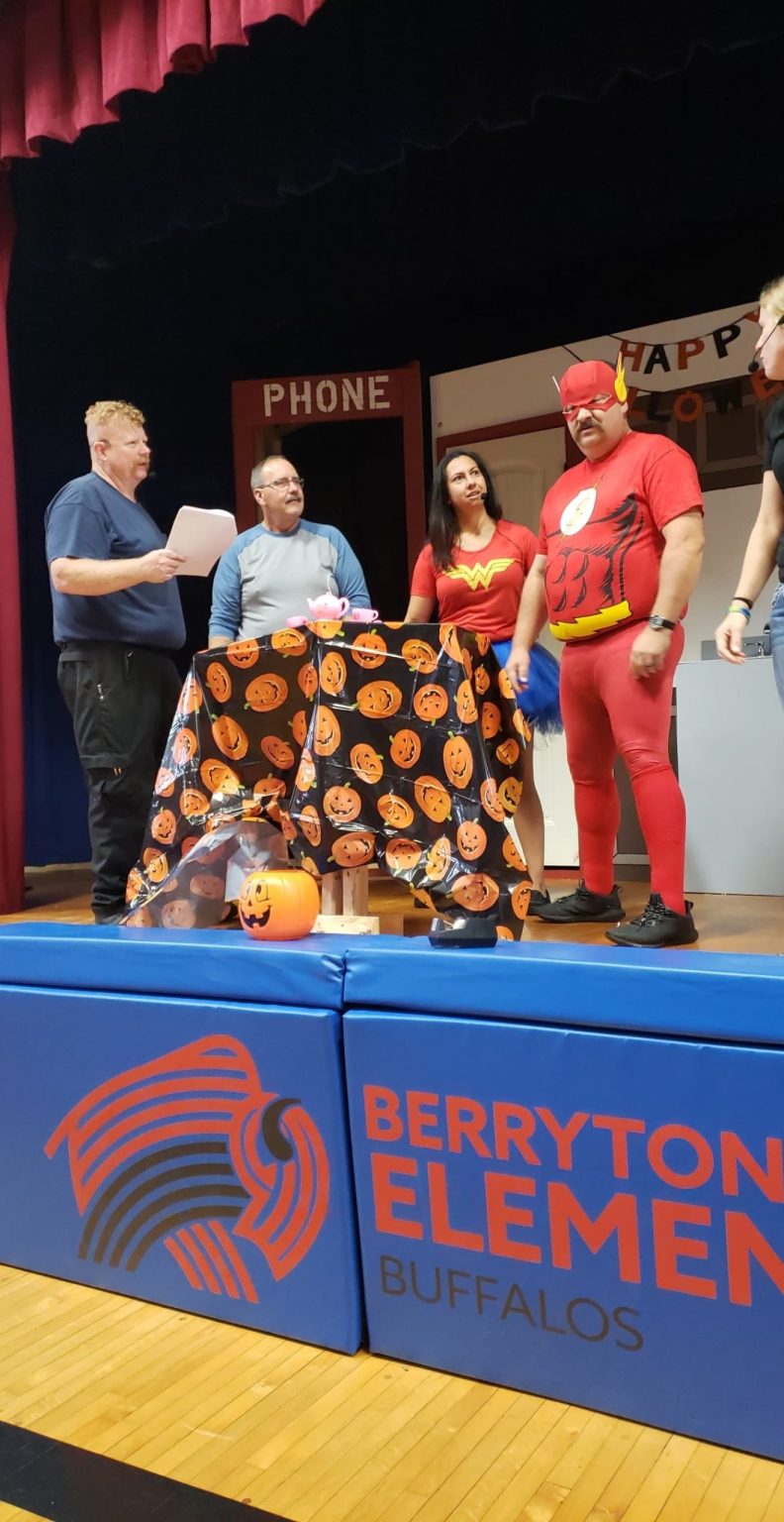 Four adults stand by a table with Halloween decorations at Berryton Elementary. Two are dressed in superhero costumes—Wonder Woman and The Flash—while two others wear regular clothes and hold papers.
