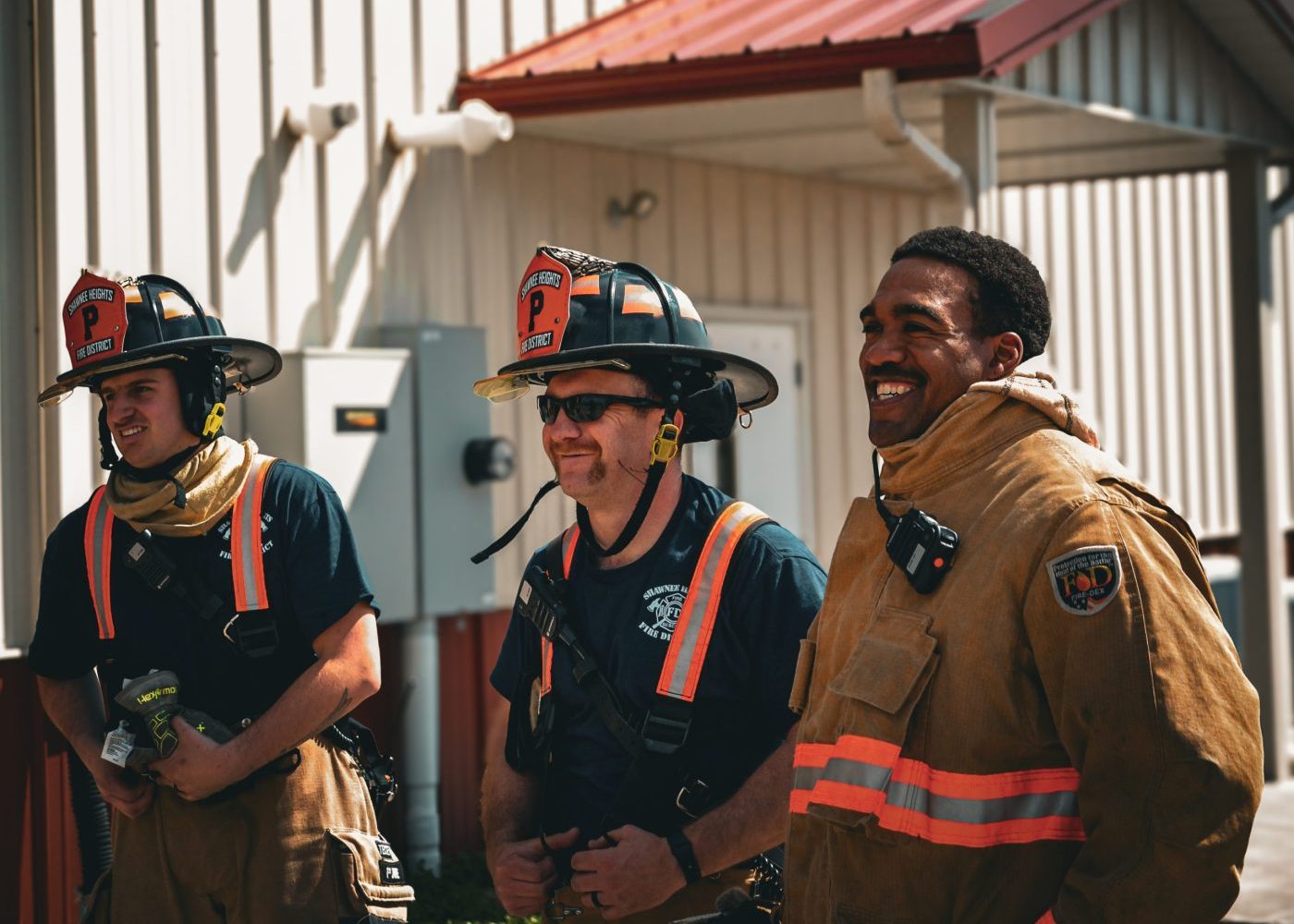 Three firefighters stand together outside a building, smiling. Two wear black shirts and firefighter helmets with gear, while one wears a tan fire jacket. The background shows a metal building with a red roof.