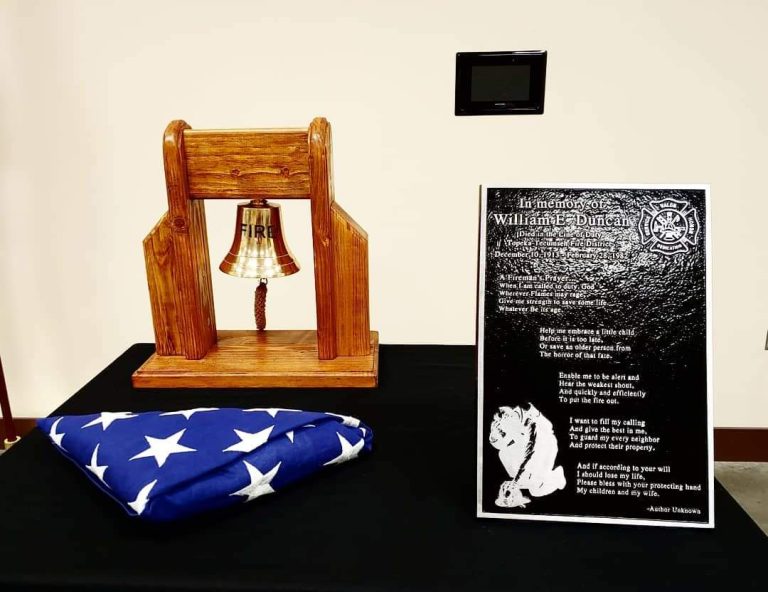 A folded American flag, a wooden bell stand marked "FIRE," and a memorial plaque rest on a black table, honoring William T. Duncan. The plaque features a firefighter’s emblem and a tribute message.