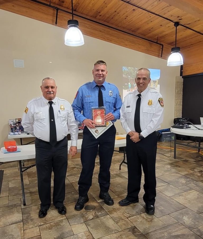 Three men in fire department uniforms stand indoors; the man in the center, wearing a blue shirt, holds a red plaque and certificate while smiling. The two men beside him wear white shirts and also smile. Tables and a projector are in the background.