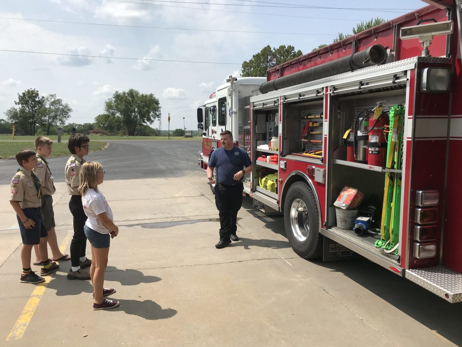 A firefighter stands beside an open fire truck, showing equipment to three boys in scout uniforms and a girl in casual clothes, all standing on a paved area near a grassy field.