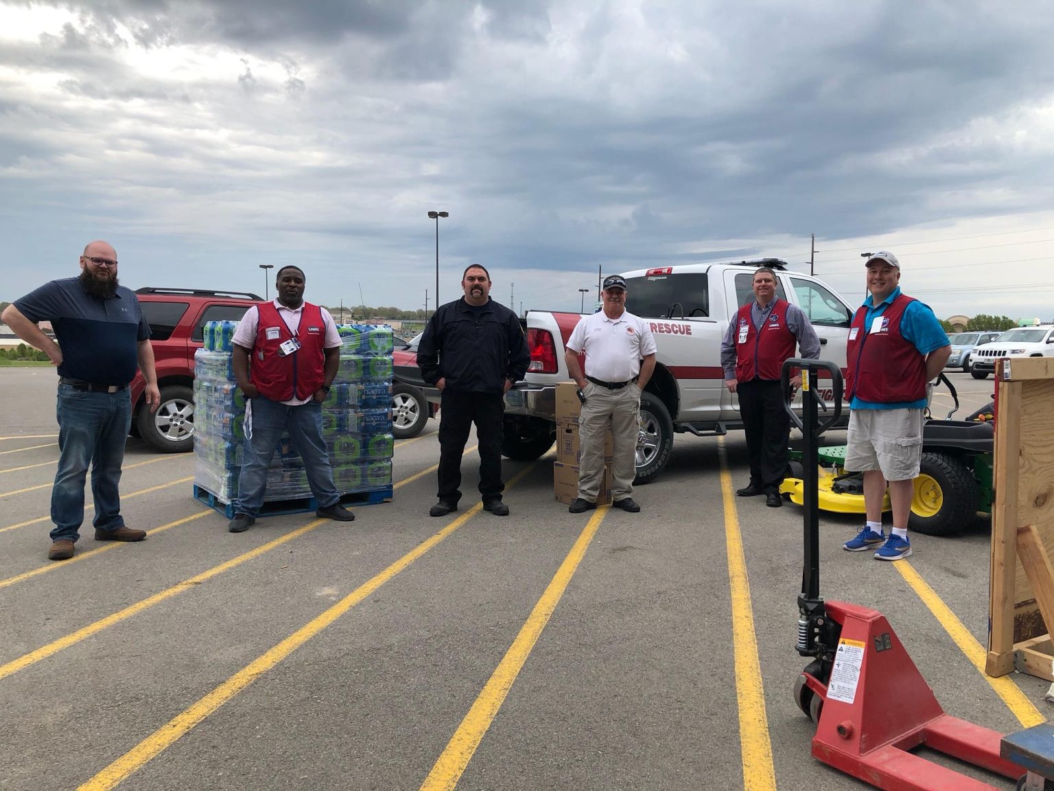 Six men stand spaced apart in a parking lot near pallets of bottled water and a rescue truck, under a cloudy sky. Some wear red vests, others are in uniforms, and carts with supplies are nearby.