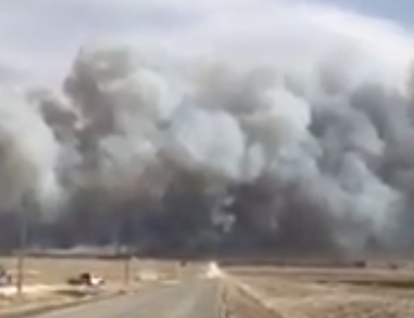 Thick clouds of smoke rise from a large wildfire burning across a flat, open field, with a road leading toward the smoke under a partly cloudy sky.