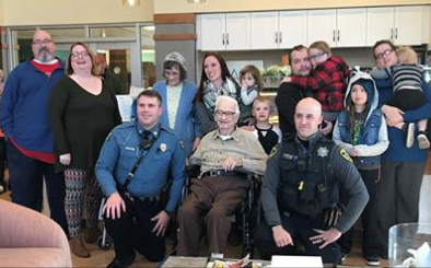 A group of people, including two police officers, several adults, children, and an elderly man in a wheelchair, pose together and smile inside a bright room with a kitchen in the background.