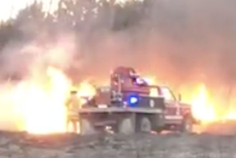 A fire truck with flashing lights is parked on dry land as firefighters work to control flames and smoke in the background, indicating an active wildfire or grass fire.