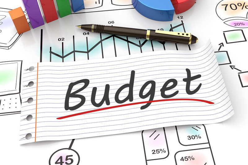 A piece of lined paper with the word "Budget" written on it lies on top of financial charts, graphs, and a pen, suggesting planning and analysis of finances.