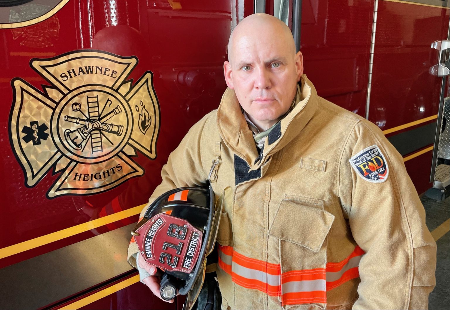 A firefighter in tan gear holds a helmet and stands next to a fire truck displaying the Shawnee Heights Fire District emblem.