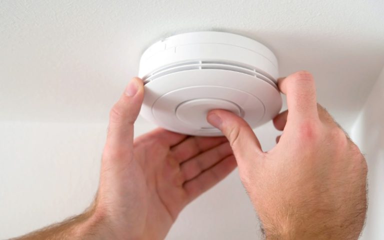 A person installs or tests a round, white smoke detector on a white ceiling using both hands.