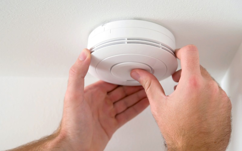 A person installs or tests a round, white smoke detector on a white ceiling using both hands.